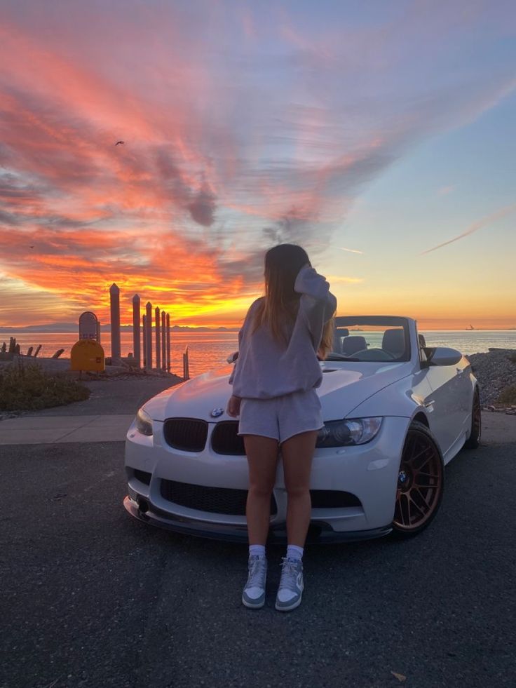 a woman leaning on the hood of a white car in front of an ocean at sunset