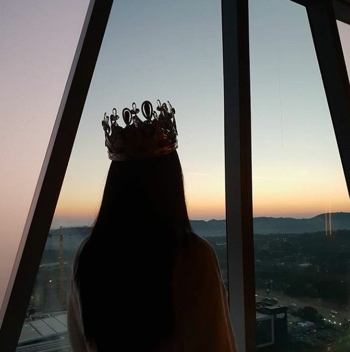 a woman wearing a crown looking out the window at the city outside her house in the evening