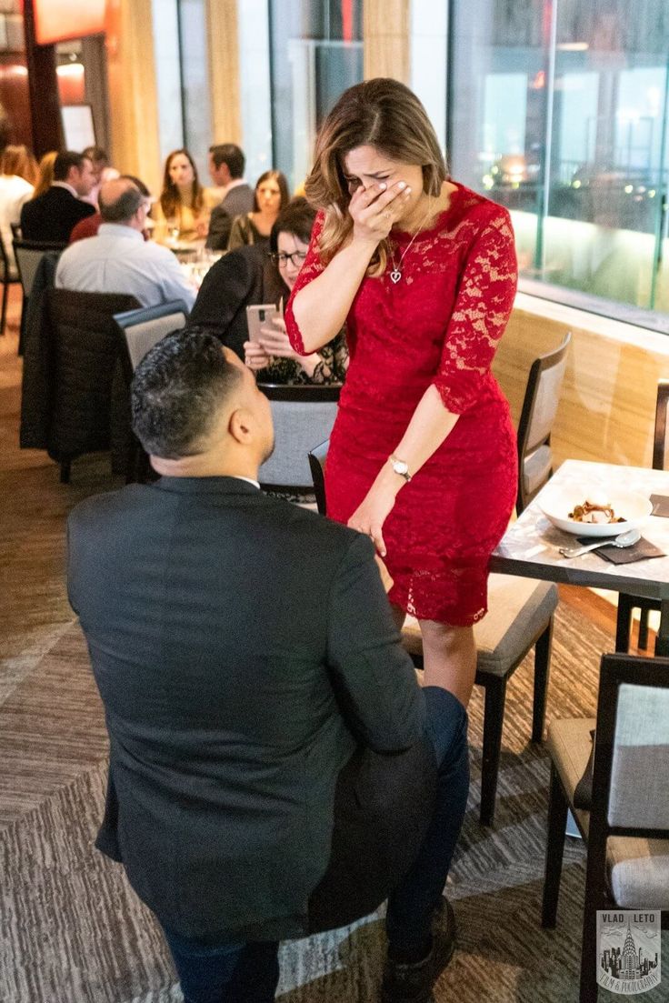 a woman in a red dress standing next to a man sitting at a restaurant table