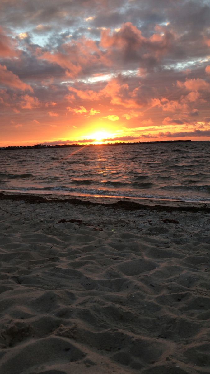 the sun is setting over the ocean with clouds in the sky and sand on the beach