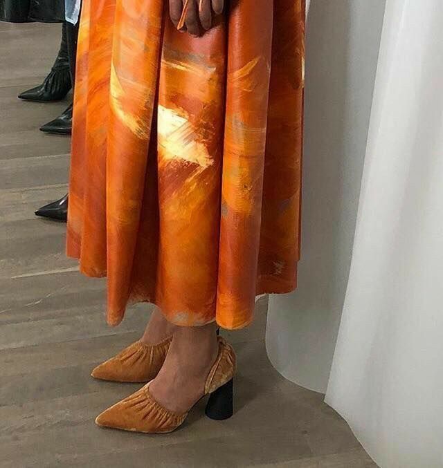 a woman standing next to a white wall with her hand on her hip wearing an orange dress