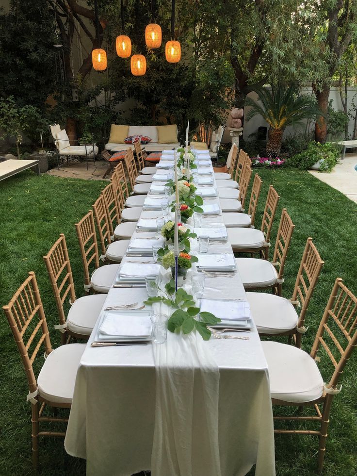 a long table is set with place settings for dinner outside in the grass, surrounded by hanging lanterns