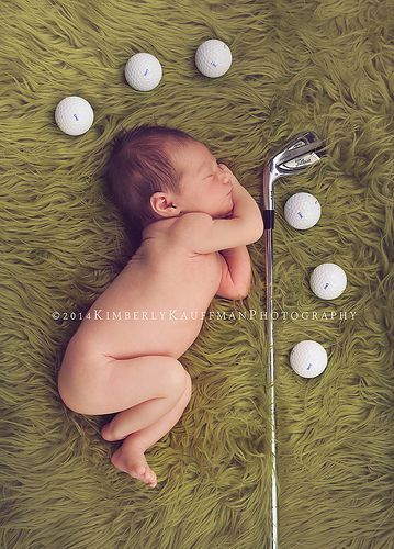 a baby laying on top of a green rug next to golf balls and a club