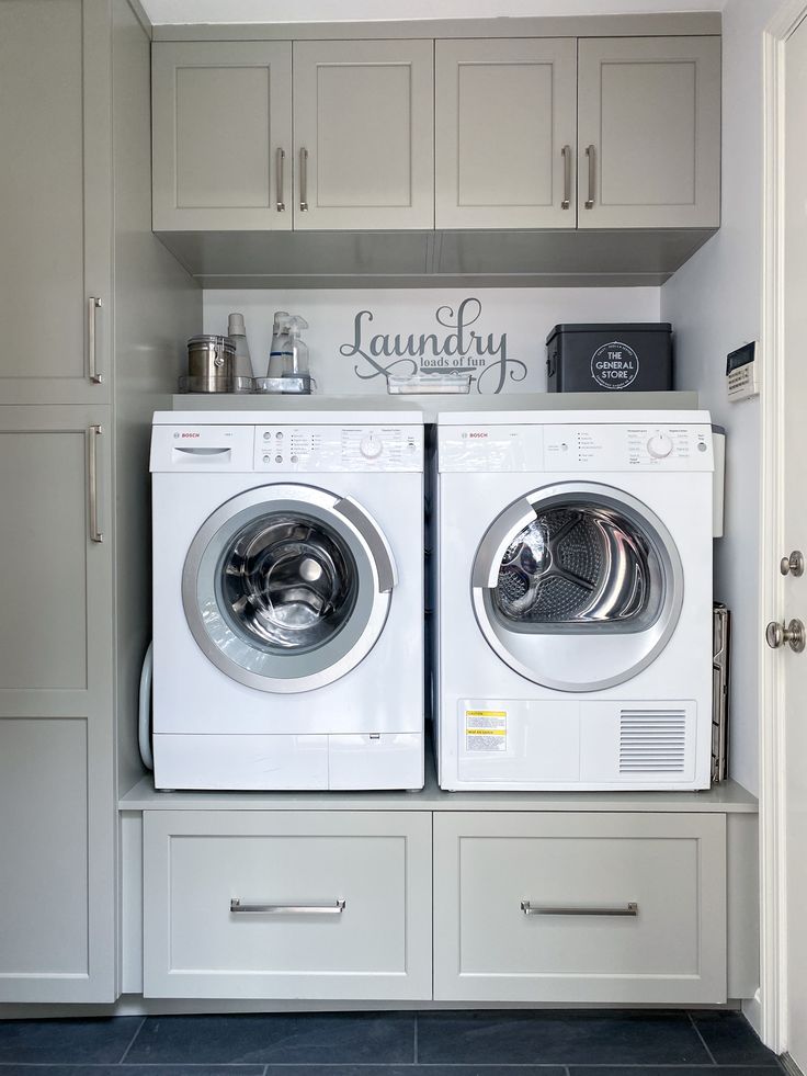 Laundry room with gray cabinetry, compact front-loading washer and dryer, and drawers below for laundry basket storage. Laundry Room With Side By Side Washer And Dryer, Basket Under Washer And Dryer, Laundry Basket Storage Under Washer And Dryer, Laundry Room Design With Front Loaders, Farmhouse Laundry Room Front Loader, Laundry Room Cabinets With Stackable Washer And Dryer, Laundry Room Design Front Loaders On Pedestals, Elevated Washer And Dryer Storage, Laundry Room With Gray Washer And Dryer