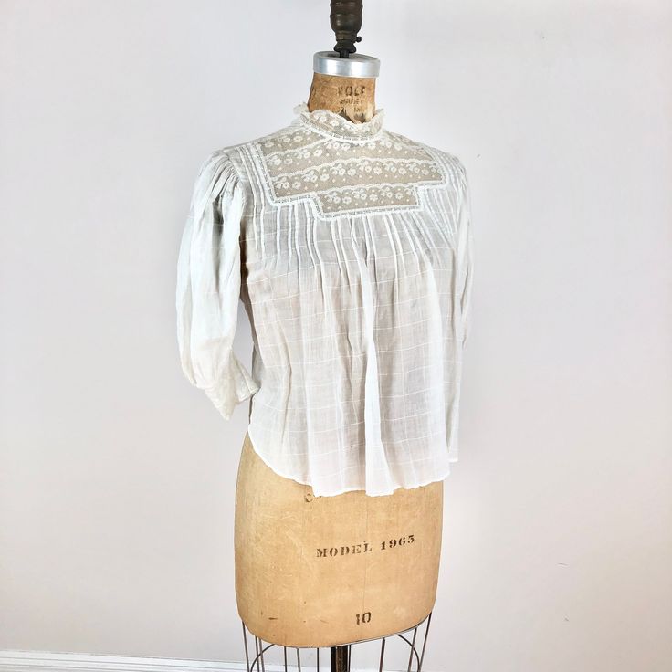 "Antique Edwardian white check cotton blouse has a lace yoke, pin tucks at the bust, and lace trimmed sleeves. The blouse has pin tuck down the length of the back, buttons for closure and a tie at the waist. Condition Overall good condition, has a stain at the back right shoulder area, tiny-small holes around the right sleeve shoulder area, a small hole near the back right pit seam, and a pink-ish stain near the shoulder. There's also a tiny hole near the back left shoulder. There's also a pink Lace Insert Blouse, Cotton Tops With Pintucks For Daywear, Cotton Peasant Top With Lace Trim For Daywear, Classic Cotton Blouse With Lace Trim, Fitted White Blouse With Pintucks, Daywear Blouse With Lace Collar, Cotton Blouse With Lace Collar For Daywear, Classic Cotton Lace Top, Fitted Cotton Peasant Top