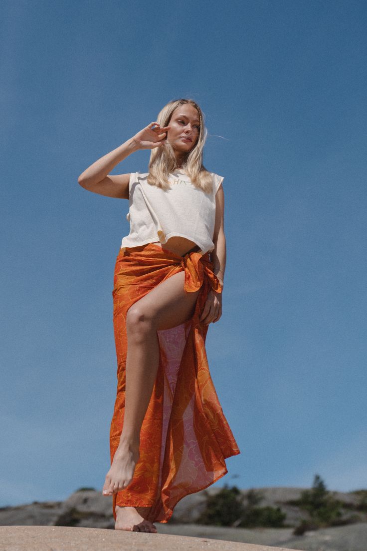 a woman standing on top of a sandy beach next to the ocean wearing an orange skirt