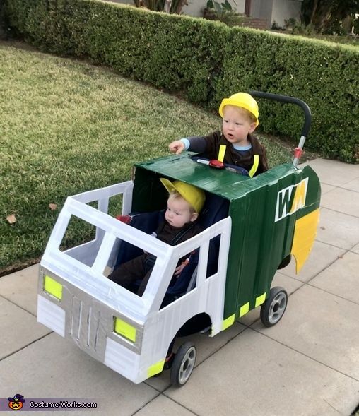 two children riding in a green and yellow toy truck on the sidewalk next to some bushes