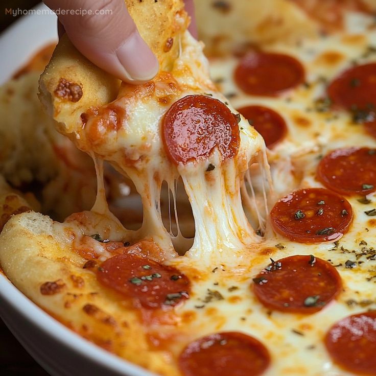 a hand holding a slice of pizza with cheese and pepperoni on it, being lifted from a white casserole dish