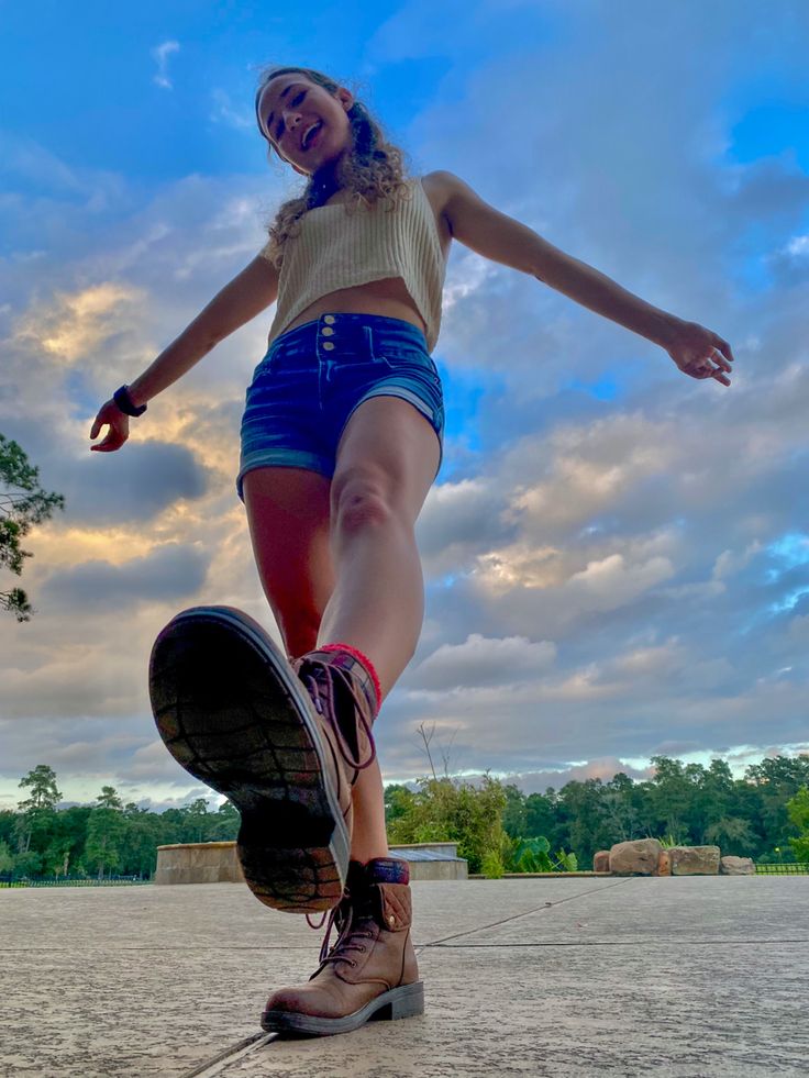 a woman in blue shorts riding a skateboard on cement ground with trees and clouds in the background