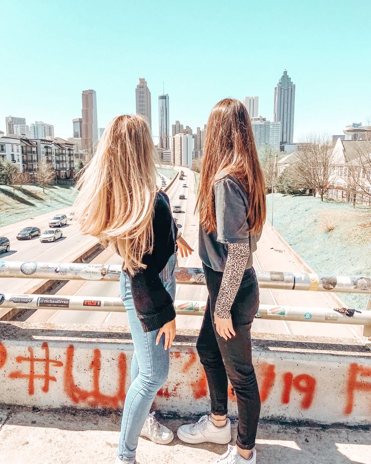 two girls standing on the edge of a bridge with graffiti written on it and buildings in the background