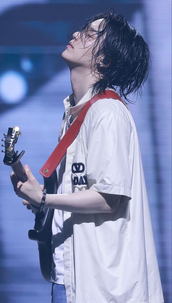 a man with long black hair holding a guitar in his right hand and wearing a red strap around his neck