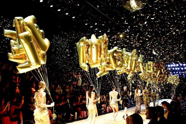 several models walk down the runway with gold balloons in the air as confetti falls on them