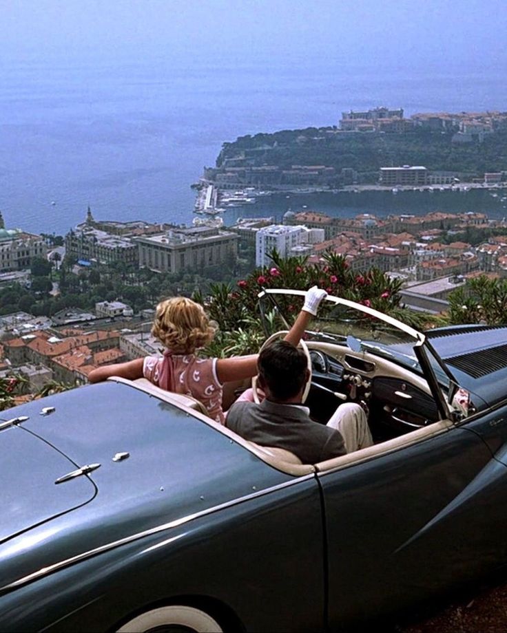 a man and woman sitting in a convertible car looking out over the city from atop a hill