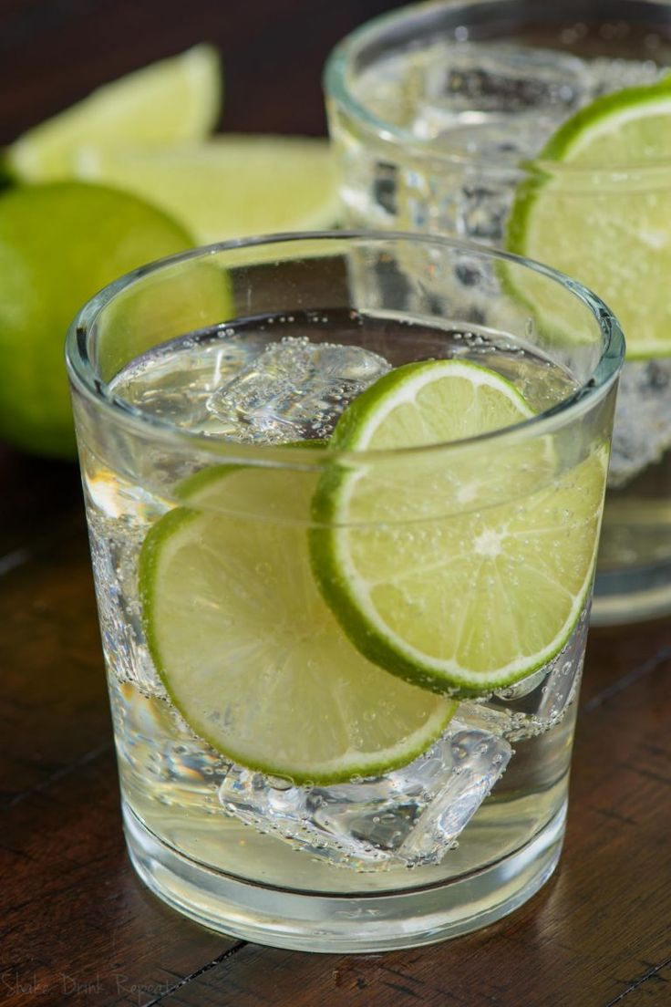 two glasses filled with gin and tonic sitting on top of a wooden table next to limes