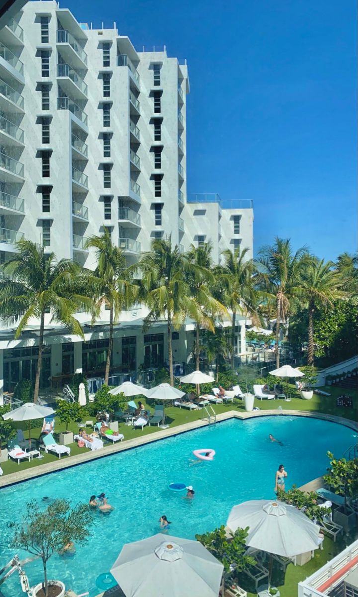 an outdoor swimming pool surrounded by palm trees and umbrellas in front of a large white building