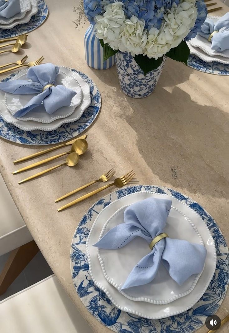 a table set with blue and white plates, gold place settings and flowers in vases