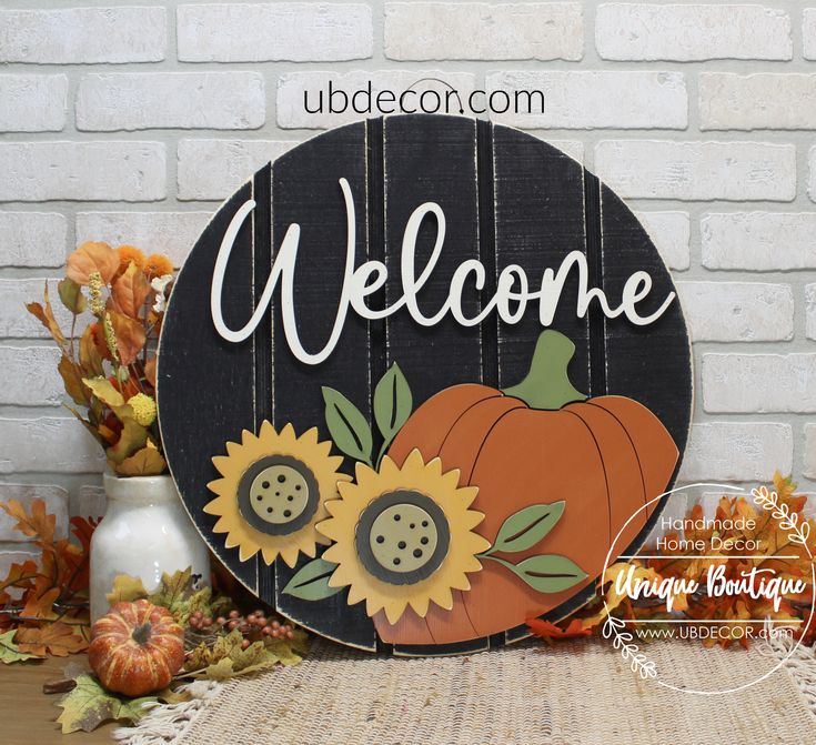 a wooden welcome sign with sunflowers and a pumpkin on the front, sitting in front of a brick wall