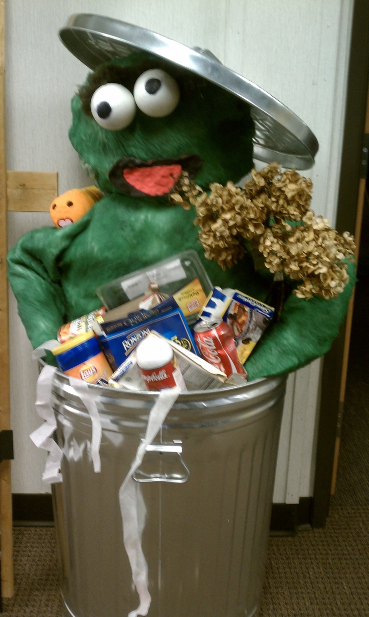 a stuffed animal sitting in a trash can filled with snacks and candy bars, while wearing a hat