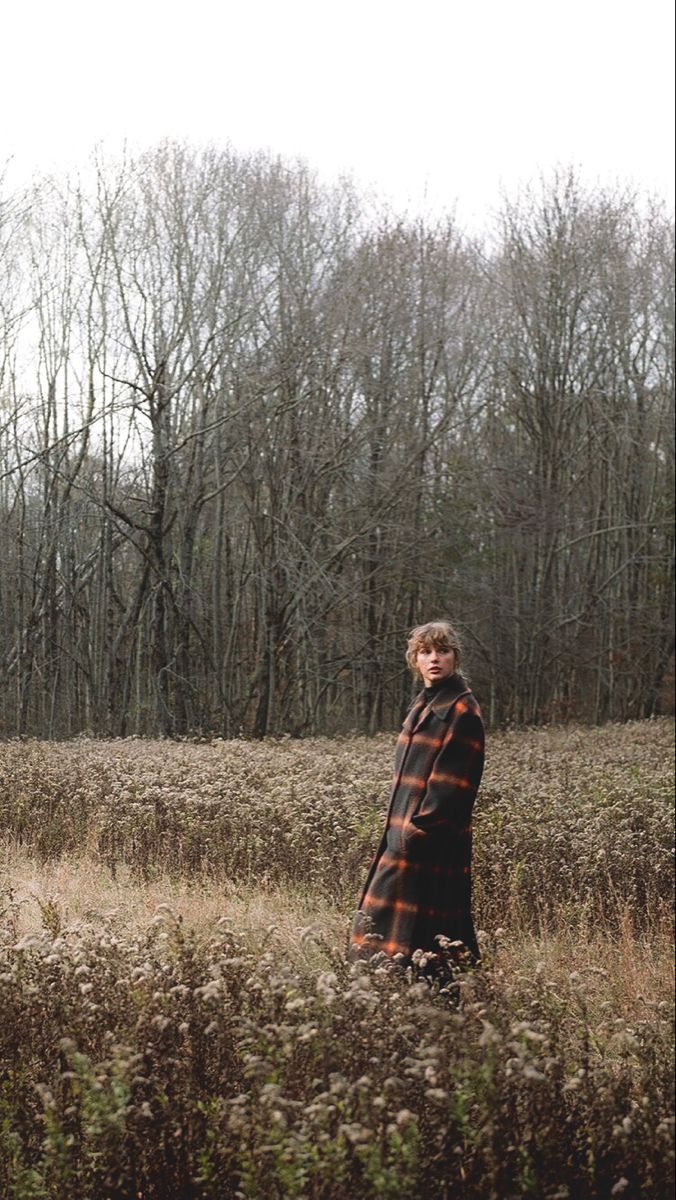 a woman standing in the middle of a field with tall grass and trees behind her