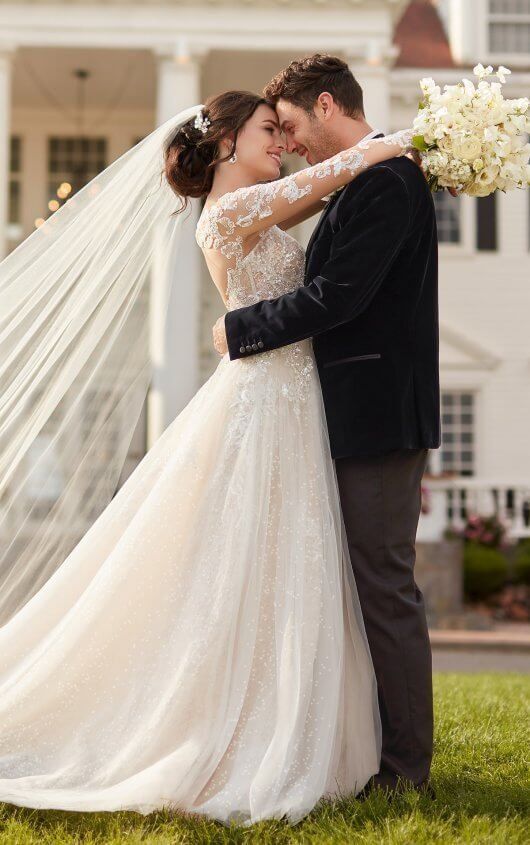 a bride and groom embracing in front of a white house with veil blowing in the wind