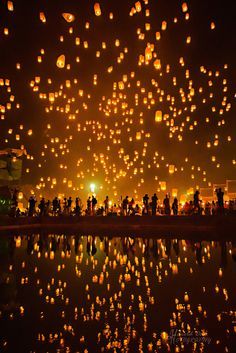 many lanterns floating in the air over water at night with people standing around and watching