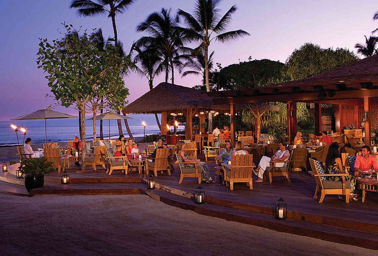 people are sitting at tables on the deck near the ocean with palm trees in the background