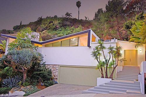 the house is surrounded by palm trees and greenery, with stairs leading up to it