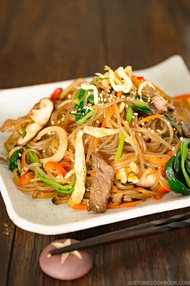 a white plate topped with noodles and vegetables next to chopsticks on a wooden table