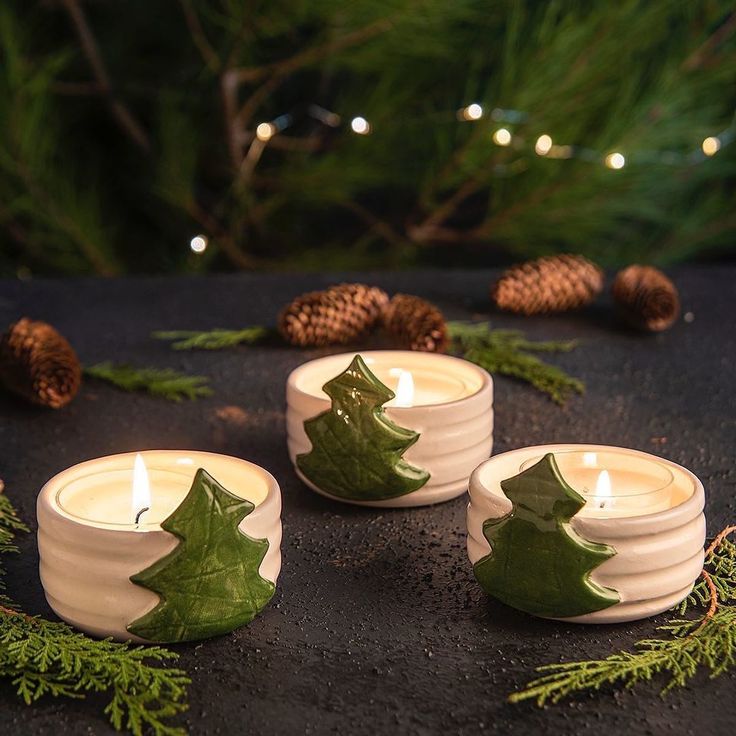 three white candles with green leaves and pine cones around them on a black surface surrounded by evergreen branches