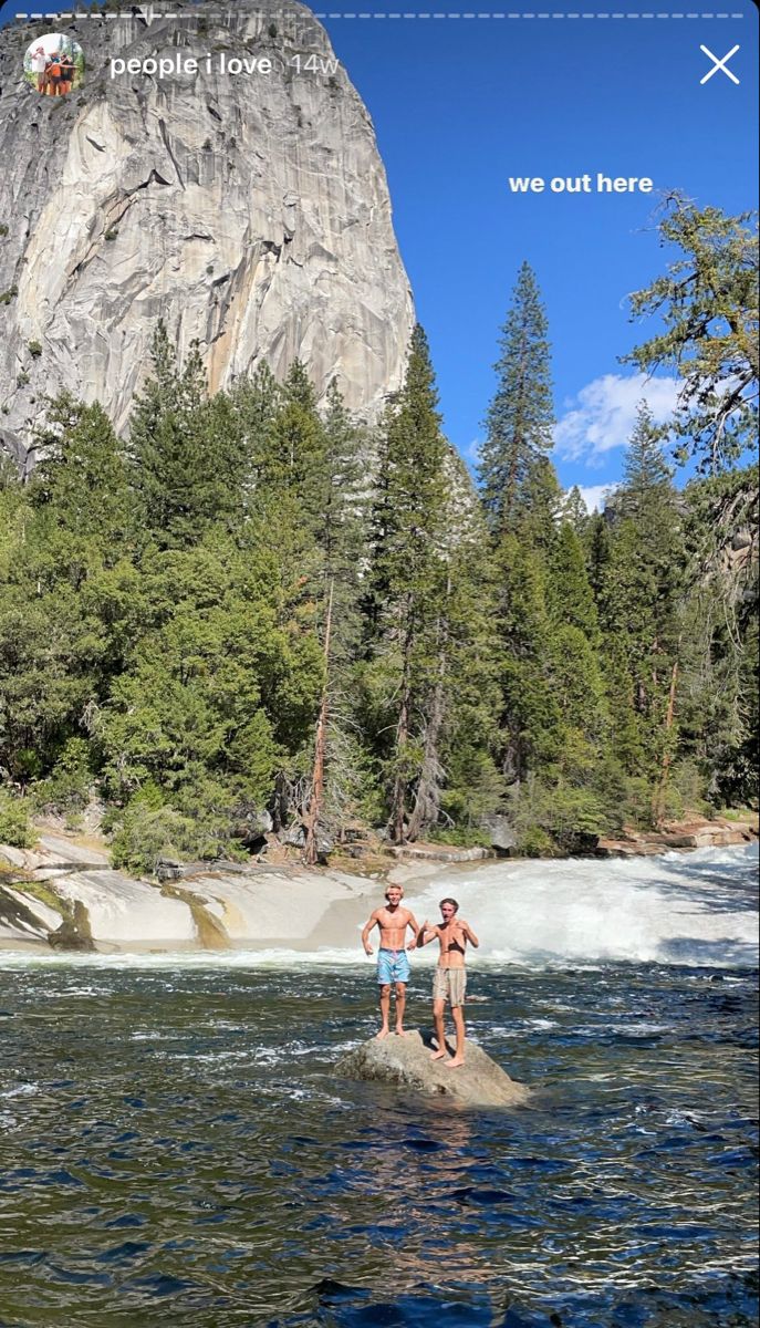 two people standing on rocks in the middle of a river with a mountain behind them