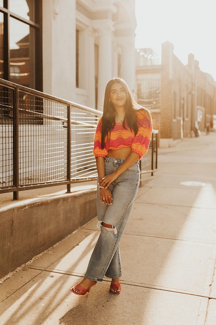 a woman standing on the sidewalk in front of a building wearing jeans and an orange top