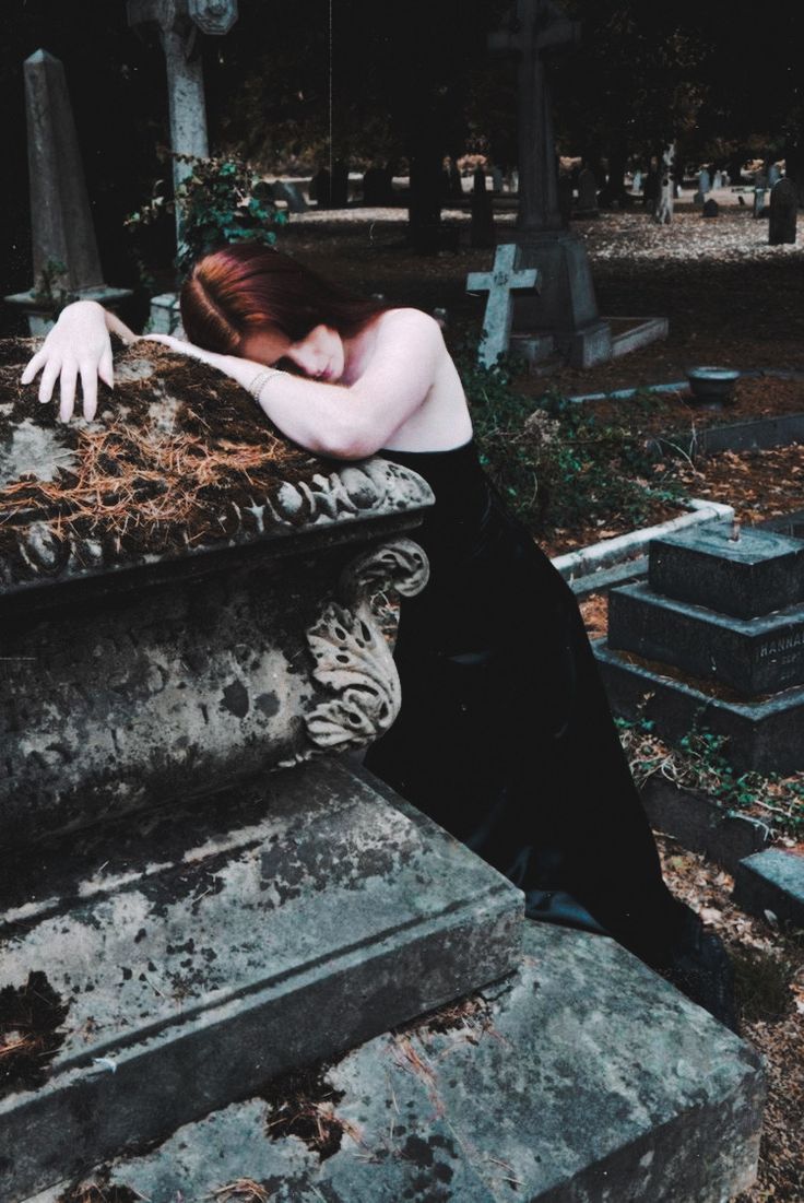 a woman with red hair is leaning on a grave in a cemetery, her hands are resting on the top of it