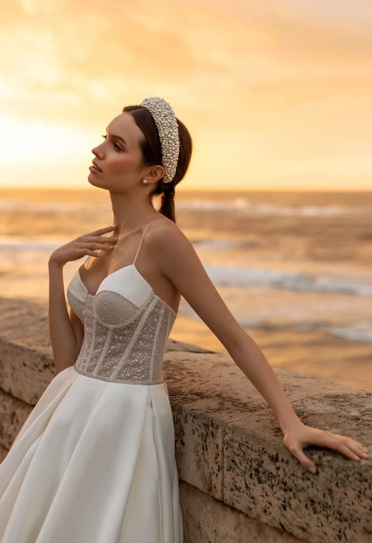 a woman in a wedding dress leaning against a wall near the ocean at sunset with her hand on her hip