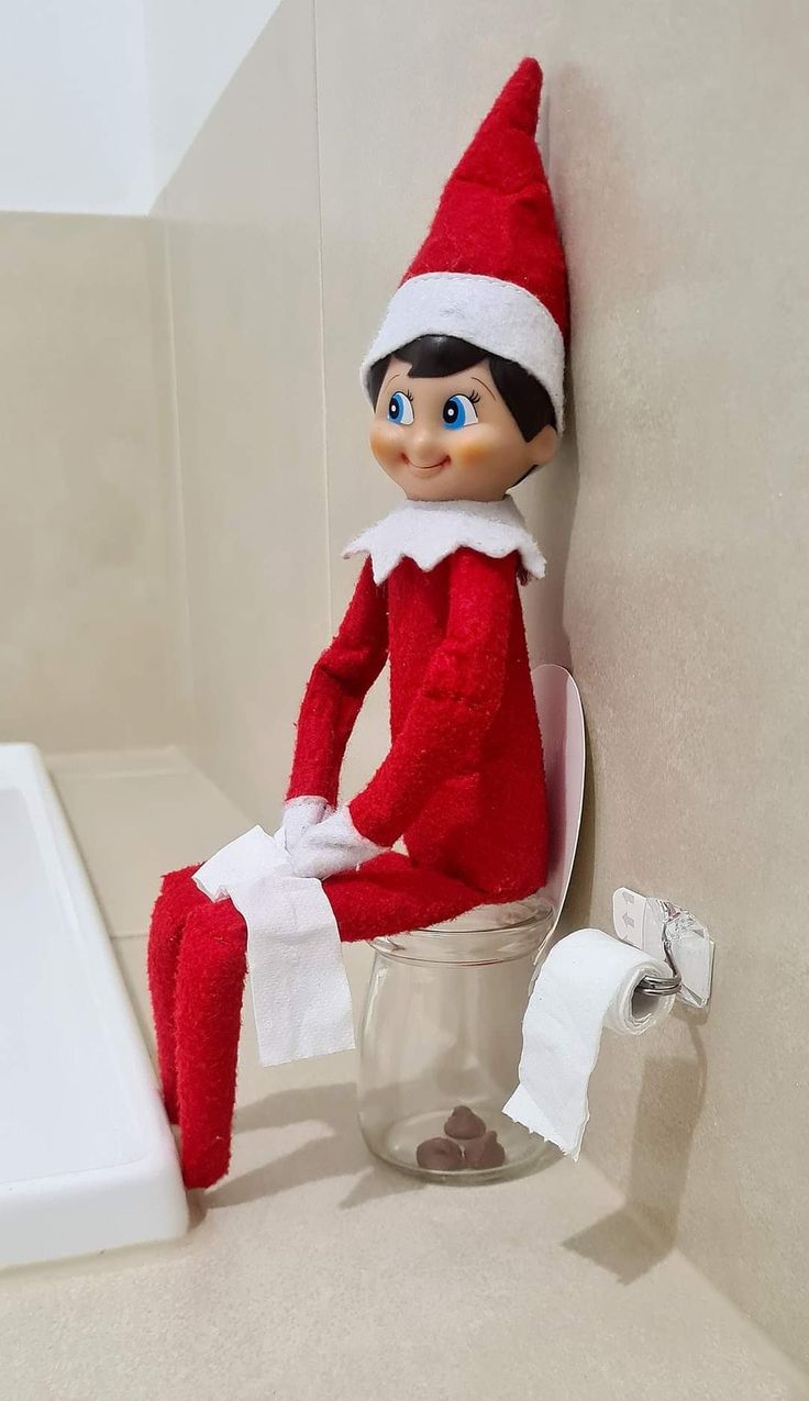 an elf sitting on top of a toilet in a bathroom next to a roll of toilet paper