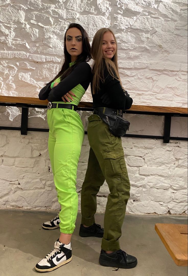 two young women standing next to each other in front of a white wall and wooden bench