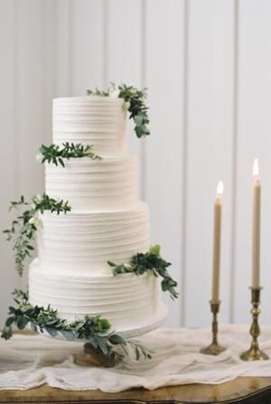 a white wedding cake with greenery on top and two candles in the back ground