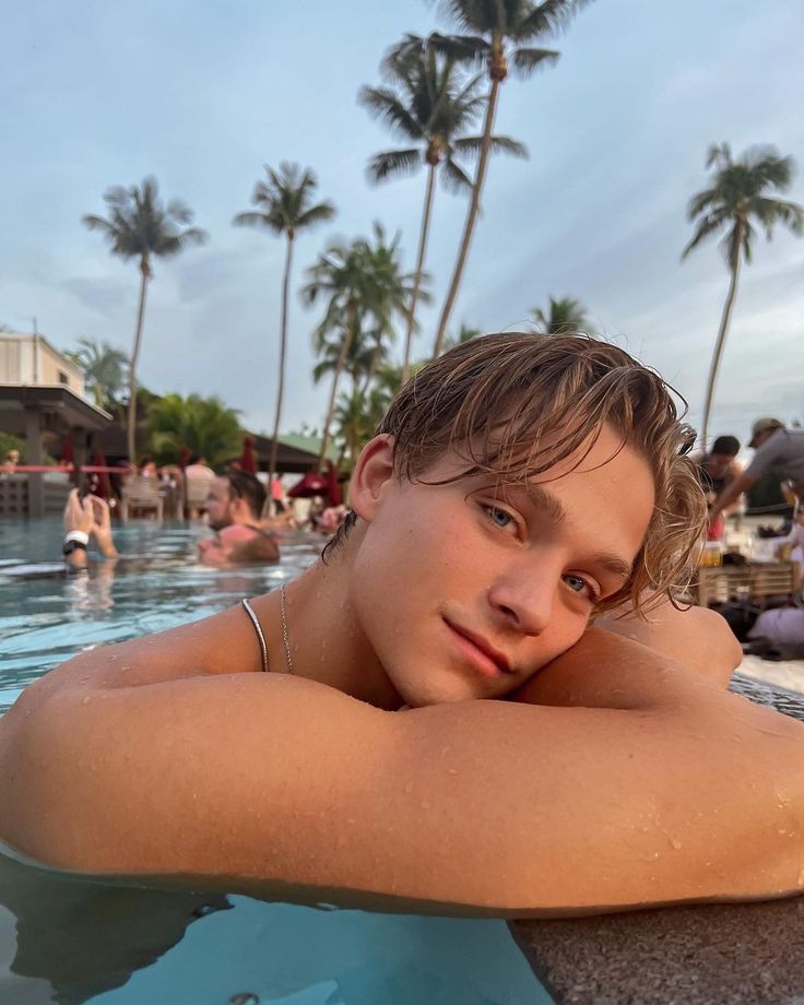 a young man sitting in the middle of a swimming pool with palm trees behind him