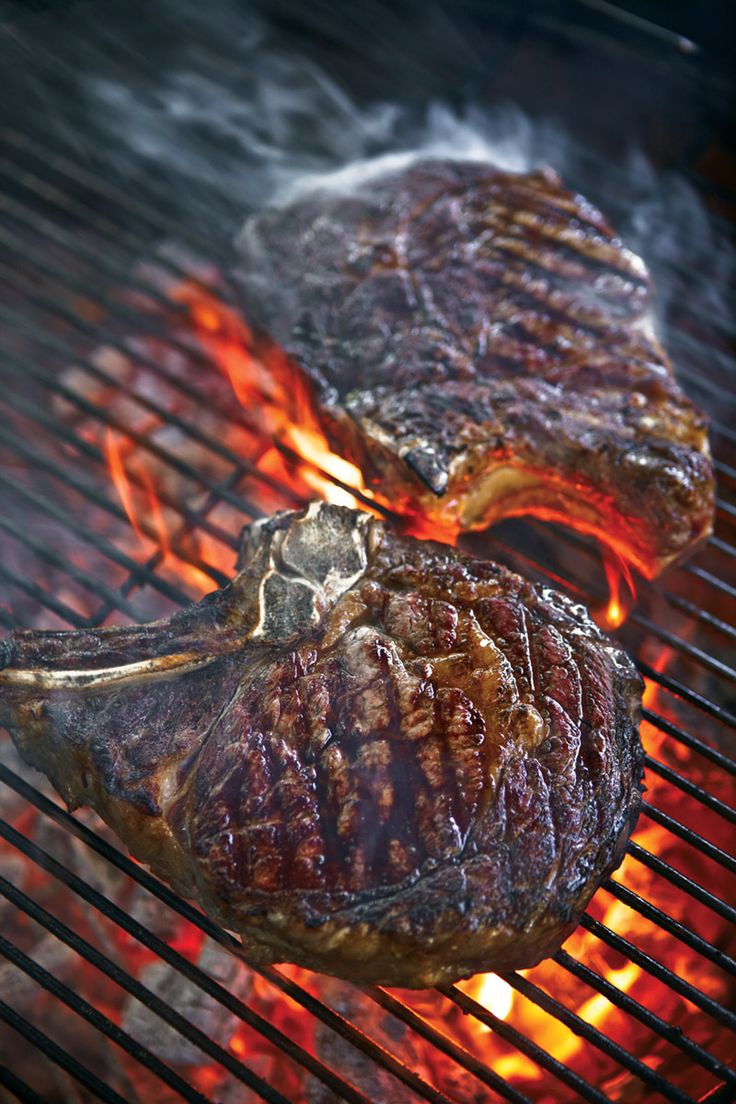 two steaks cooking on the grill with flames