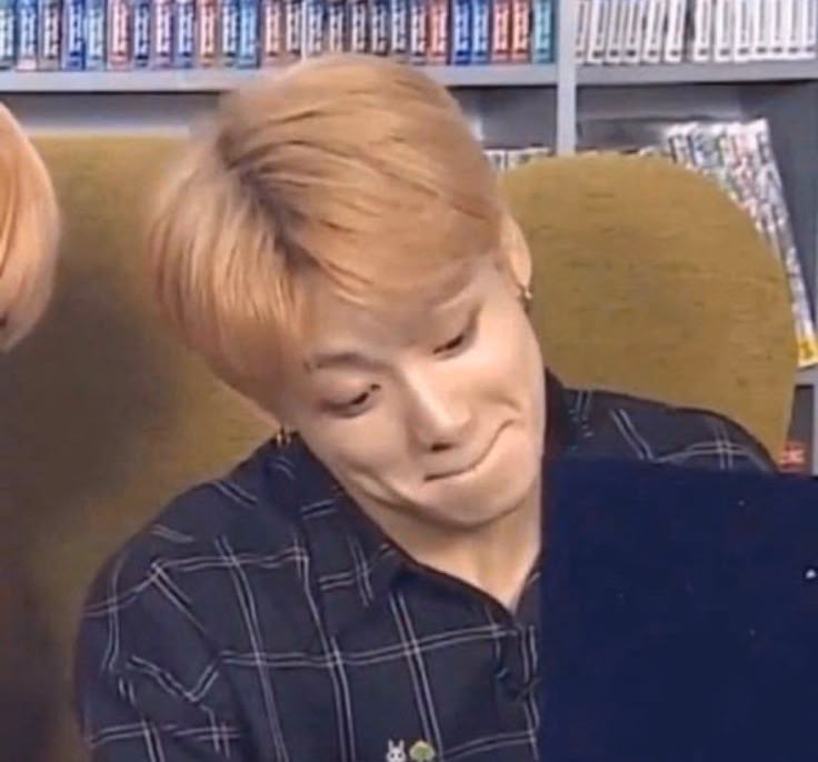 a man with blonde hair sitting in front of a book shelf and looking at his laptop