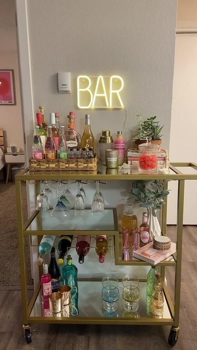 a bar cart filled with bottles and glasses on top of a hard wood floor next to a neon sign