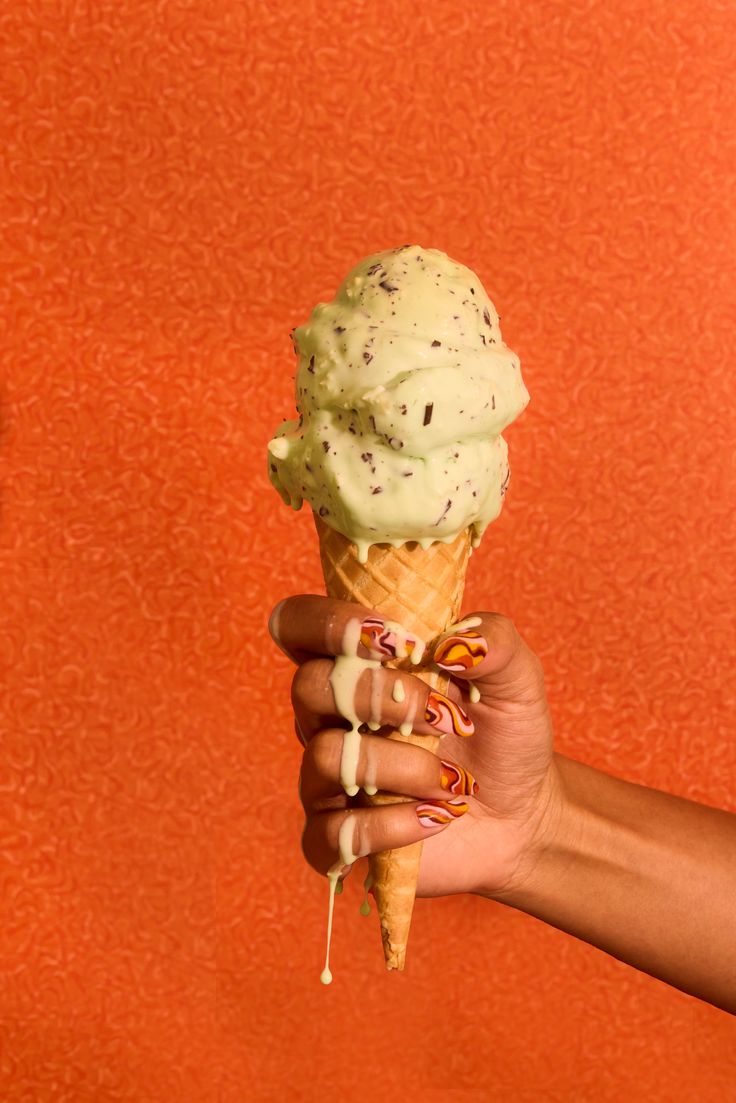 a hand holding an ice cream cone with chocolate sprinkles on orange background