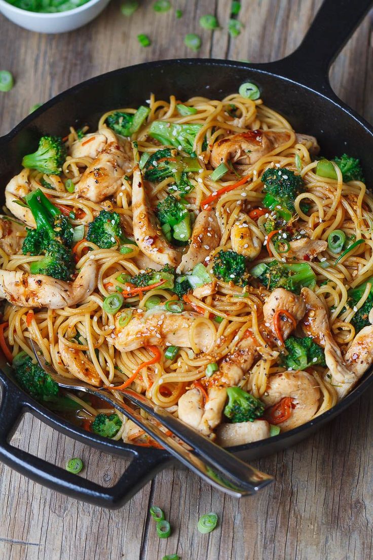 a skillet filled with noodles, broccoli and chicken on top of a wooden table