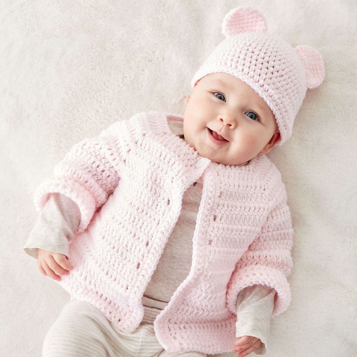 a baby wearing a pink knitted bear sweater and hat laying on a white blanket