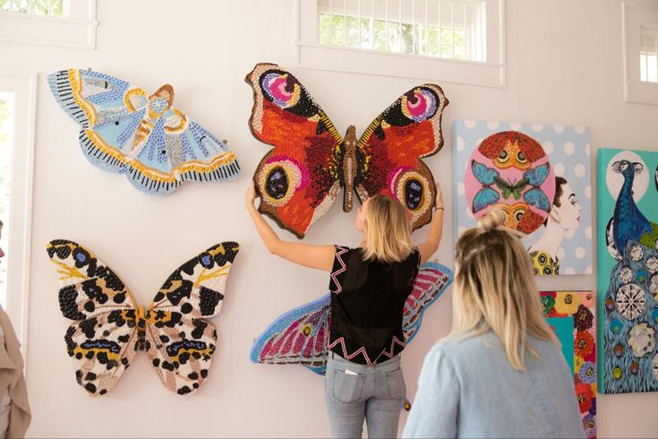 two women looking at colorful butterfly artwork on the wall in a white room with windows