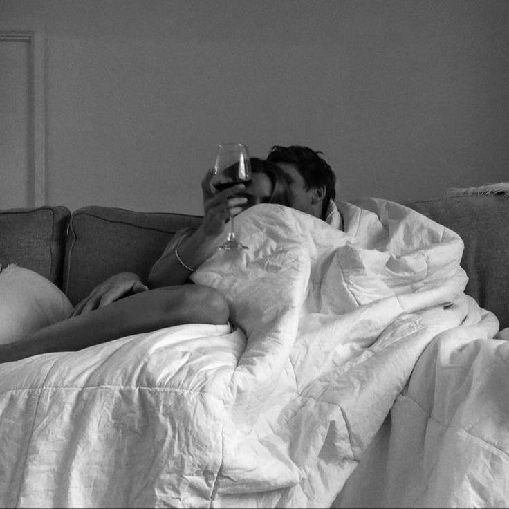 a man and woman laying on a couch under a blanket with wine glasses in their hands