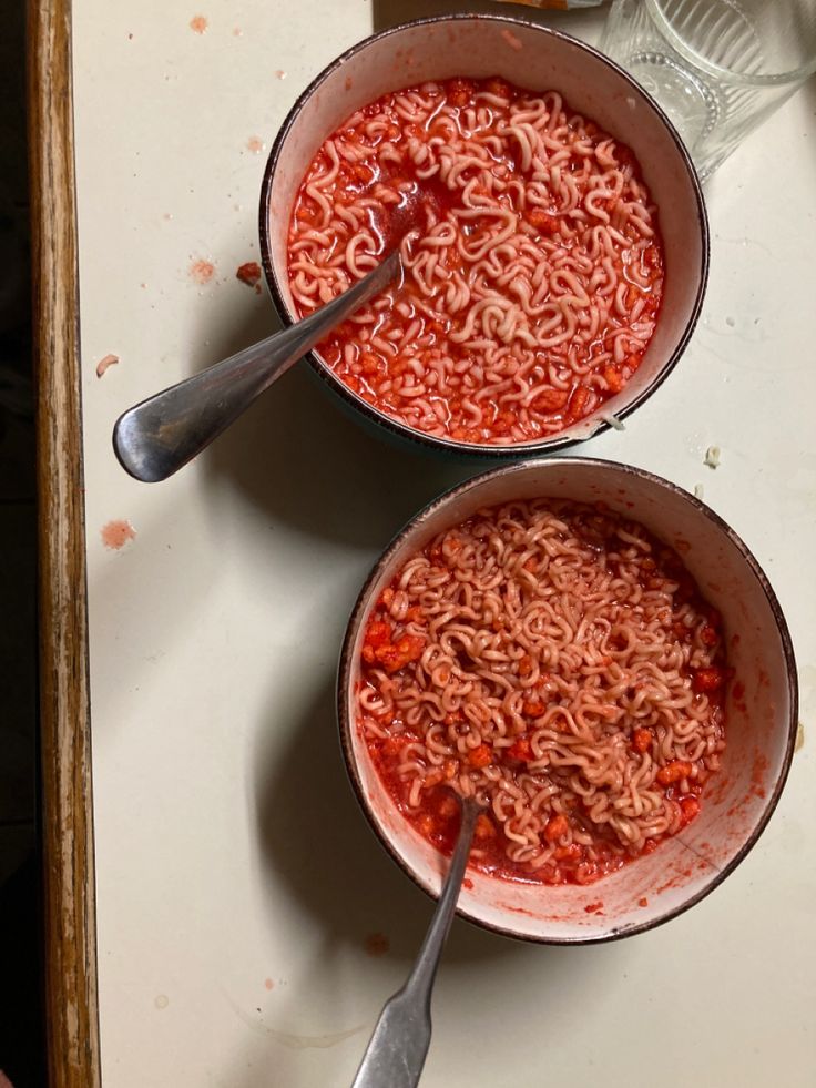 two bowls filled with noodles on top of a white counter next to silver spoons