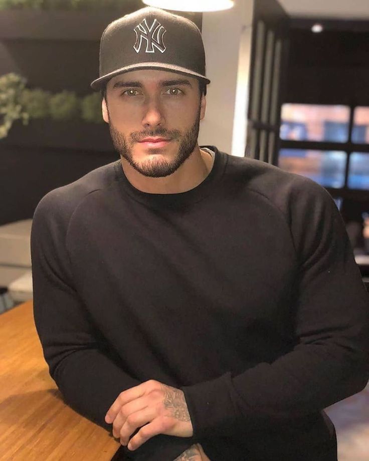 a man wearing a new york yankees hat sitting in front of a wooden table with his arms crossed