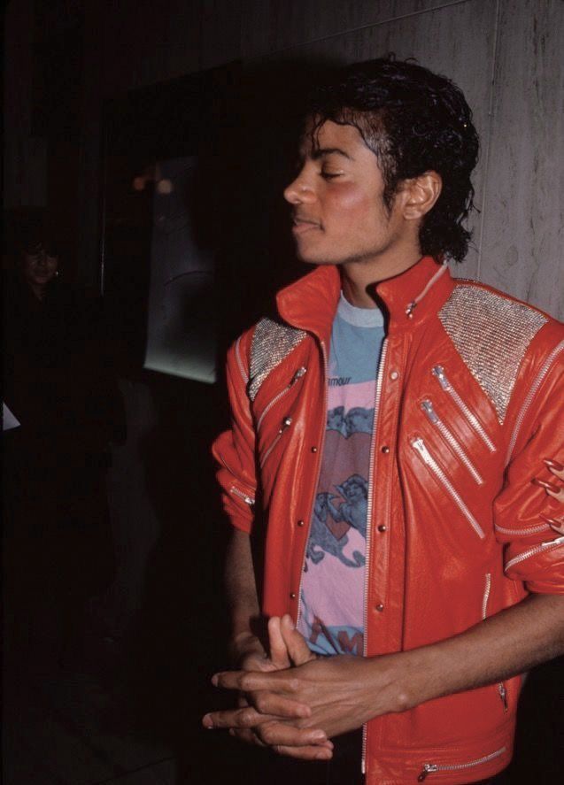 a man in an orange leather jacket standing next to a wall with his eyes closed