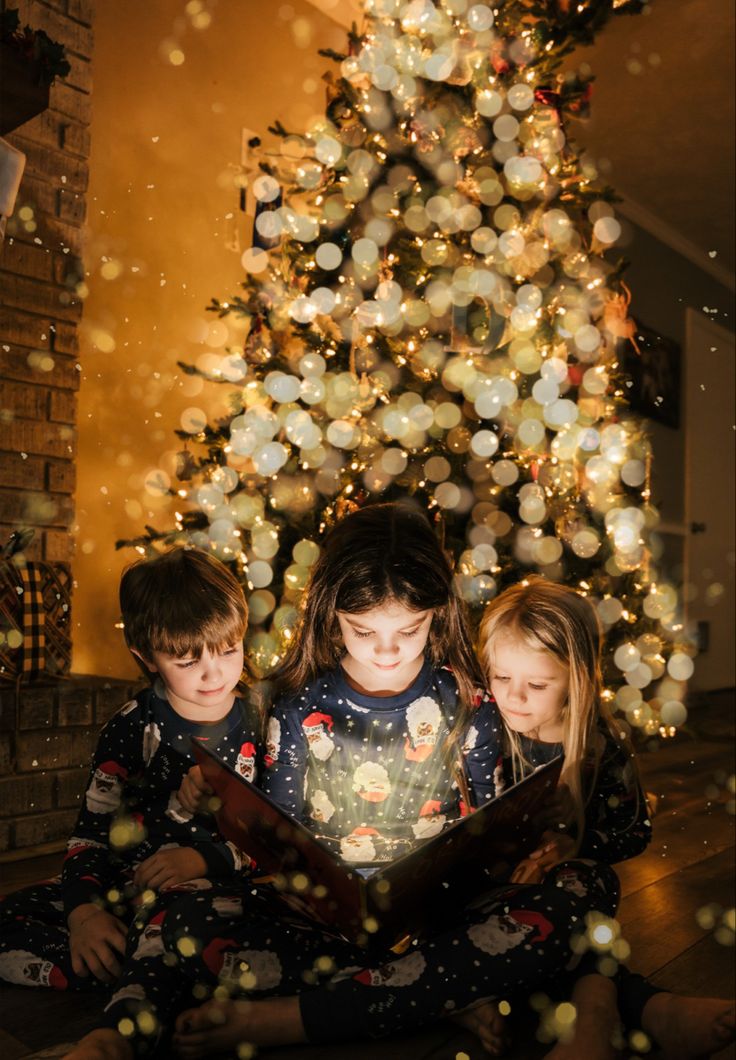 three young children sitting in front of a christmas tree looking at an open book together