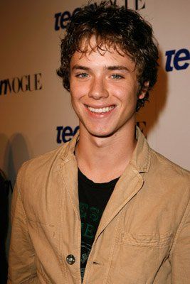 a young man smiling at the camera with his hands in his pockets and wearing a tan jacket